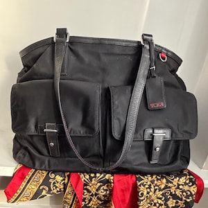 May include: Black tote bag with two front pockets, leather straps, and silver-tone hardware. The bag has a rectangular shape and a tag with the brand name. A red and gold patterned scarf is visible at the bottom.