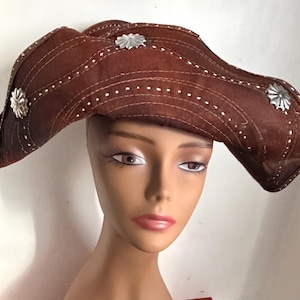 May include: A brown hat with a wide brim, featuring white stitching and silver-colored star-shaped embellishments. The hat is displayed on a mannequin head. The hat's design includes a series of parallel stitched lines.