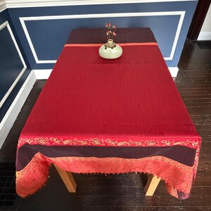 May include: A rectangular red tablecloth with a decorative border. The tablecloth has a dark burgundy top section and a decorative border with gold accents. A small, light green vase with a floral arrangement sits on top of the tablecloth.