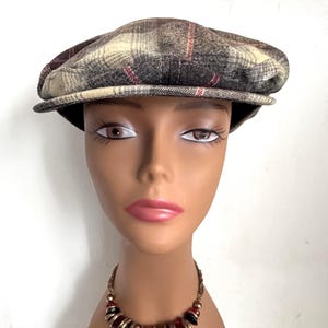 May include: A plaid newsboy cap in shades of brown, beige, and burgundy. The cap has a rounded crown and a small, stiff brim. The hat is displayed on a mannequin head.