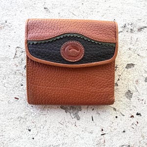 May include: A brown leather wallet with a flap closure. The wallet features a textured surface and a decorative black and green accent on the flap. A circular logo is visible on the flap.