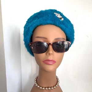 May include: A mannequin head wearing a teal fuzzy hat with a jeweled embellishment, tortoiseshell sunglasses, and a pearl necklace. The hat is a vibrant teal color, and the sunglasses have dark lenses.