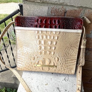May include: A beige crossbody bag with a crocodile-like texture. The top section is a deep burgundy, and the bag has a long, adjustable strap. A small gold-toned metal plate with the brand name is visible.