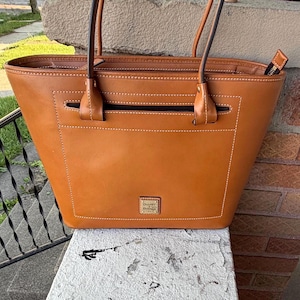 May include: A brown leather tote bag with two handles and a rectangular shape. The bag has a front pocket with a zipper and a small logo on the bottom. The stitching is white, and the bag appears to be new.