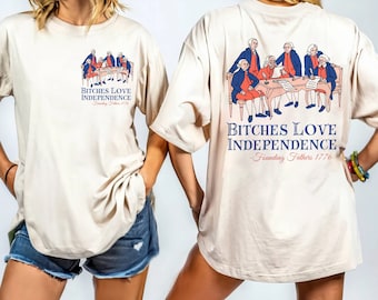May include: A beige t-shirt featuring a graphic of the Founding Fathers seated around a table. The text on the shirt reads "Bitches Love Independence - Founding Fathers 1776".