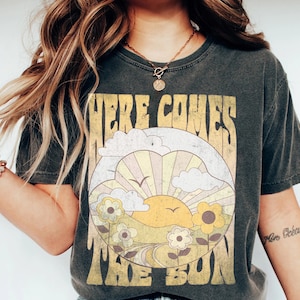 May include: A dark gray t-shirt with a vintage-style graphic of a sun rising over a field of flowers. The text "Here Comes The Sun" is printed in a retro font.