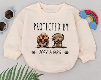 May include: A cream-colored sweatshirt with the text "PROTECTED BY" and two dogs, a golden retriever and a poodle, with the names "ZOEY & PARIS" printed below.