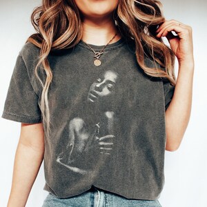 May include: A dark gray t-shirt with a graphic print of a woman's face. The shirt has a relaxed fit and a crew neckline. The woman is wearing a gold necklace with a coin pendant.