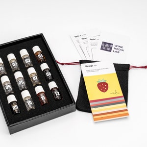 May include: A black box with 12 small glass bottles labeled with numbers 1 through 12. The bottles are arranged in four rows of three. The box is on a white surface. There is a small black bag with a red drawstring and a card with a strawberry on it. The card says "WINE MEDIA LAB" and "Orange Test".
