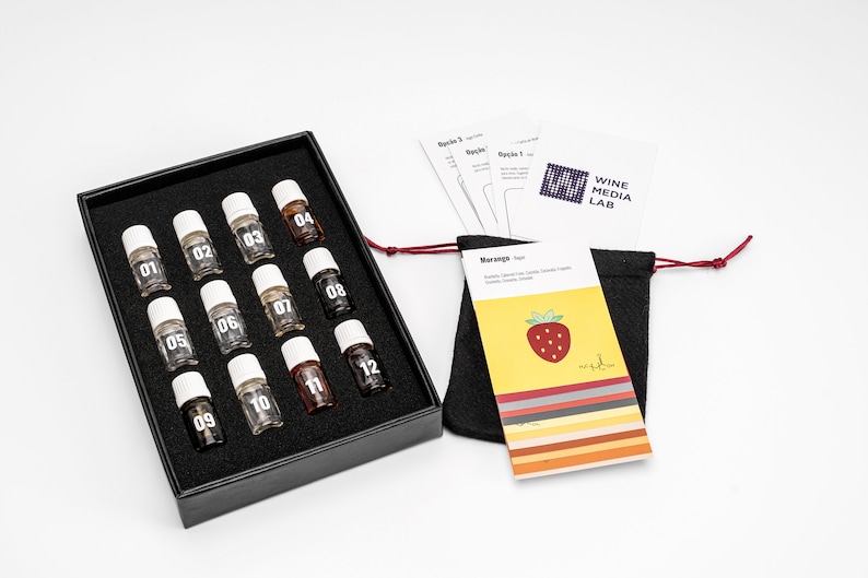 May include: A black box with 12 small glass bottles labeled with numbers 1 through 12. The bottles are arranged in four rows of three. The box is on a white surface. There is a small black bag with a red drawstring and a card with a strawberry on it. The card says "WINE MEDIA LAB" and "Orange Test".