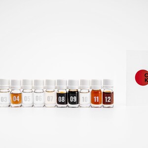 May include: Twelve small glass bottles with white caps, numbered 01 to 12, containing different colored liquids. A white card with a red circle and the text "COR KY." is to the right of the bottles.