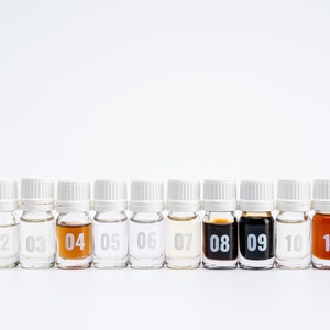 May include: Twelve small glass bottles with white caps, lined up in a row. Each bottle is labeled with a number from 01 to 12. The bottles contain different colored liquids, including clear, light brown, and dark brown.