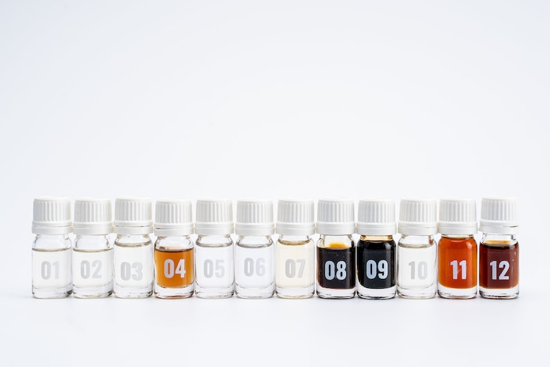 May include: Twelve small glass bottles with white caps, lined up in a row. Each bottle is labeled with a number from 01 to 12. The bottles contain different colored liquids, including clear, light brown, and dark brown.