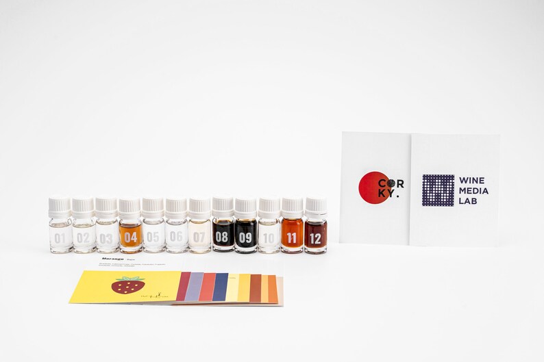May include: Twelve small glass bottles with white caps, numbered 1 through 12, containing different colored liquids. The bottles are arranged in a row on a white surface. A card with the text "COR KY." and a red circle is visible in the background. Another card with the text "WINE MEDIA LAB" is also visible in the background.