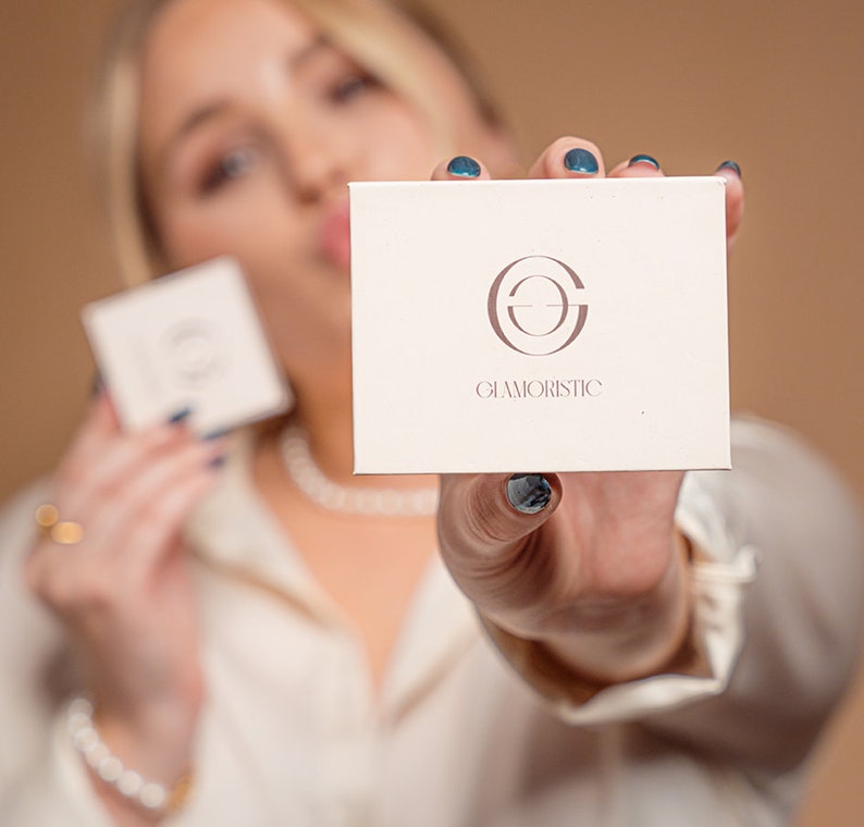 May include: A white box with a logo of a circle with a line through it and the text "GLAMORISTIC" below. The box is being held by a person with blue nail polish.