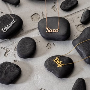 May include: Three gold necklaces with personalised names on black stones. The names are "Chiara", "Sara", and "Lily".