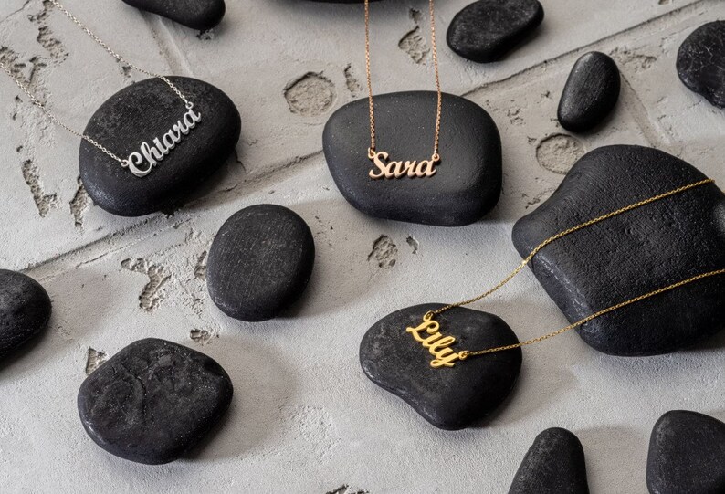 May include: Three gold necklaces with personalised names on black stones. The names are "Chiara", "Sara", and "Lily".