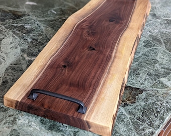 Puede incluir: Una bandeja de servir de madera grande con un borde vivo y dos asas de metal negro. La bandeja está hecha de madera de nogal y tiene un acabado natural.