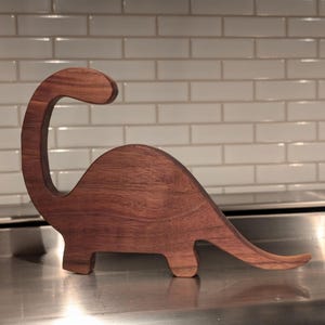 May include: A wooden dinosaur sculpture, carved from dark brown wood. The dinosaur is standing on a silver surface.