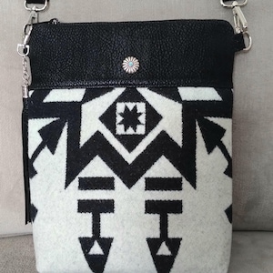 May include: Black leather crossbody bag with a white and black geometric patterned flap. The bag has a silver chain strap and a silver concho.