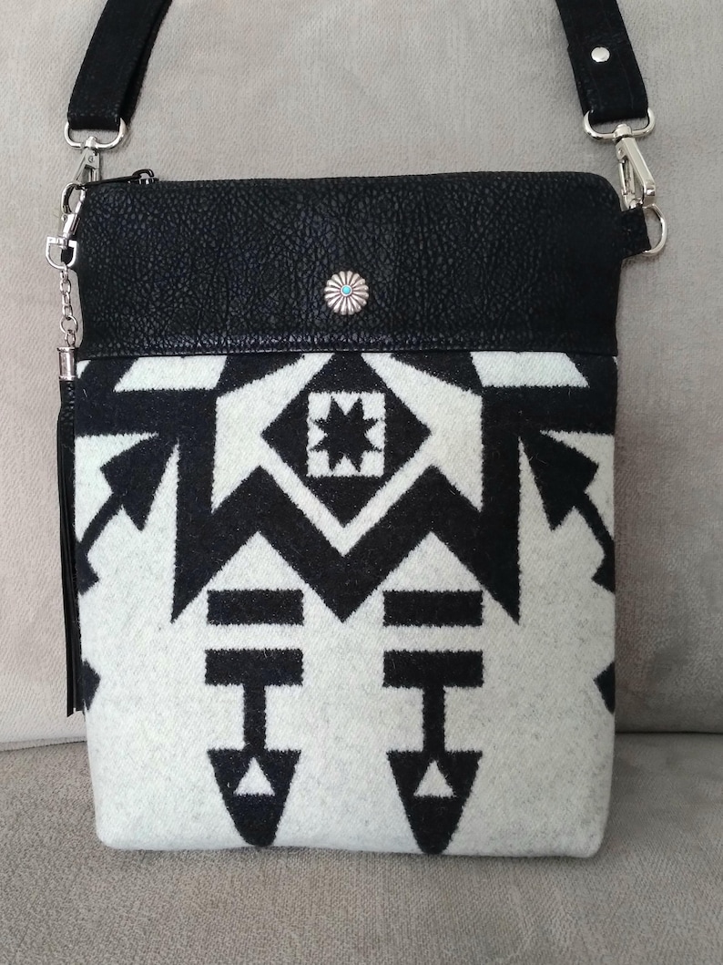 May include: Black leather crossbody bag with a white and black geometric patterned flap. The bag has a silver chain strap and a silver concho.