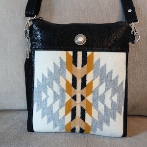 May include: Black leather crossbody bag with a geometric patterned front panel in white, gray, gold, and black. The bag has a silver-tone clasp and a black adjustable strap. A decorative silver button with a white center is on the front.