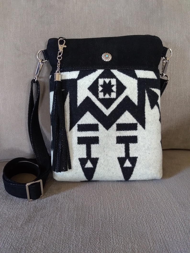 May include: Black and white geometric patterned crossbody bag with a black leather strap and tassel. The bag has a black suede flap with a turquoise stone embellishment.