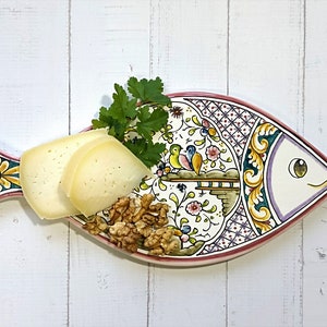 Pode incluir: Um prato de servir de cerâmica branca em forma de peixe com um design floral colorido. O prato é decorado com flores e pássaros rosa, amarelos, verdes e azuis. O prato está cheio de queijo e nozes.