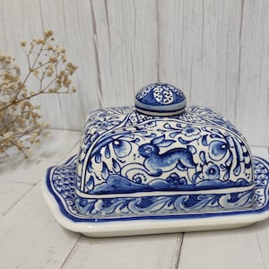 May include: A rectangular ceramic butter dish with a lid featuring hand-painted blue floral patterns and a rabbit motif. The dish has a matching blue and white decorative border on the base and a small round knob on top of the lid for easy handling.