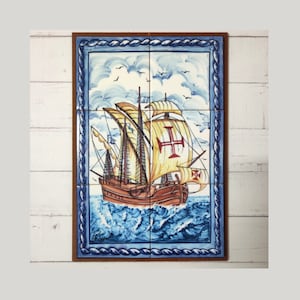 Pode incluir: Um azulejo de cerâmica pintado à mão que representa uma caravela portuguesa navegando num mar azul com nuvens brancas. O navio tem uma cruz vermelha na sua vela e está rodeado por ondas azuis. O azulejo está emoldurado numa moldura de madeira castanho com um desenho de corda azul e branca.
