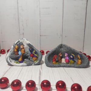 Pode incluir: Duas cenas de natividade de cerâmica cinza com pequenas figuras de Maria, José e o menino Jesus. As figuras estão vestidas com roupas coloridas. As cenas estão sobre um fundo de madeira branca.