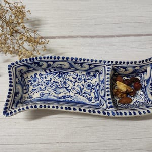 May include: A white ceramic serving dish with a blue and white floral design. The dish is divided into two sections. One section has a blue and white floral design and the other section has a blue and white floral design with a small amount of nuts in it.