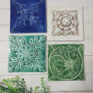 May include: Four square ceramic tiles with embossed designs in shades of blue, white, and green. The tiles feature a floral pattern, a stylized flower, and an abstract design. The tiles are displayed on a white wooden surface.