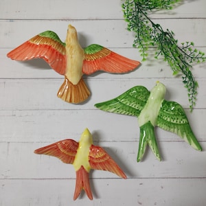 May include: Three ceramic bird ornaments with outstretched wings. The birds are painted in shades of orange, green, yellow, and brown. They are set against a white wooden backdrop, with a green plant visible in the background.