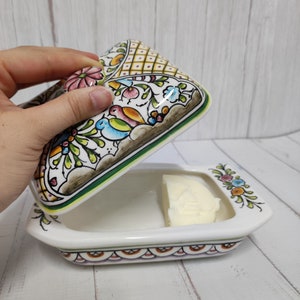 Puede incluir: Un mantequillero de cer&aacute;mica blanca con un dise&ntilde;o floral en tonos verdes, azules, amarillos, rosas y marrones. La tapa est&aacute; abierta y hay una barra de mantequilla dentro.