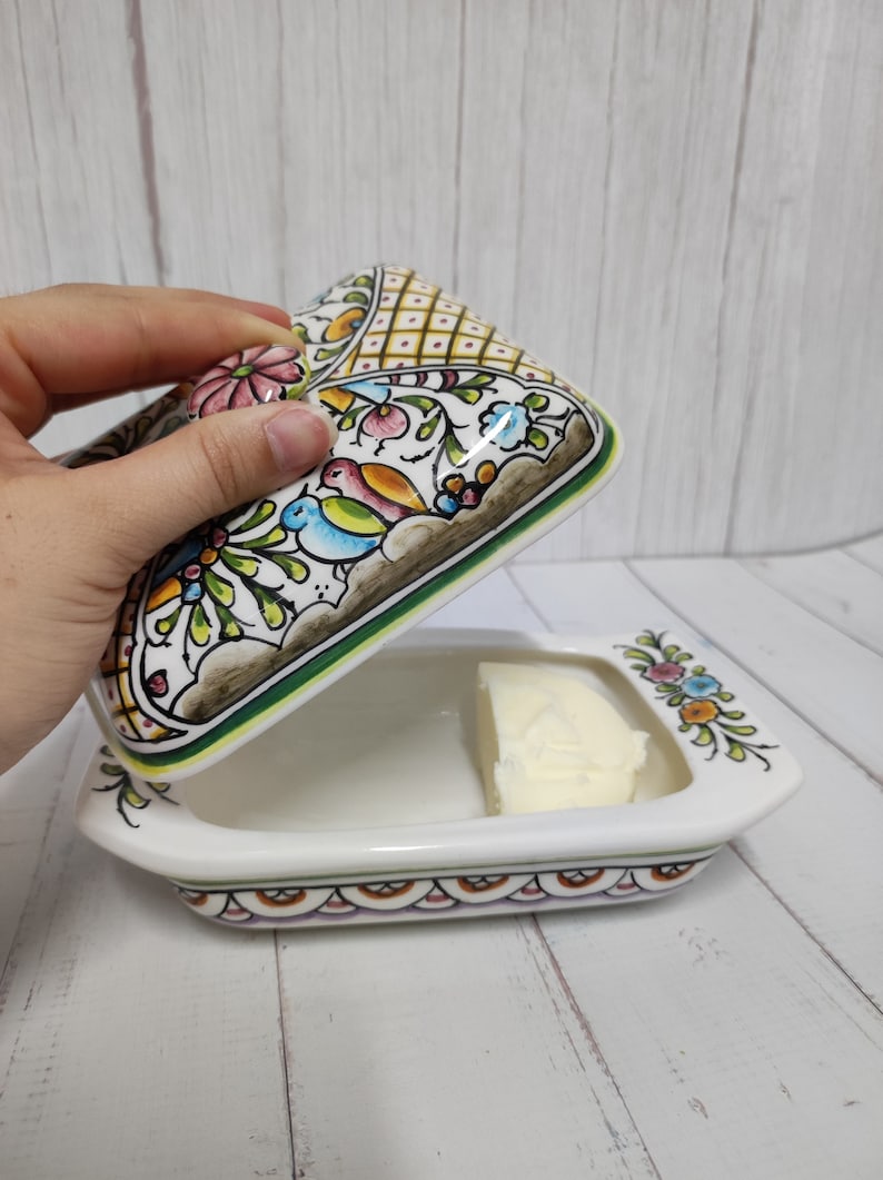 Puede incluir: Un mantequillero de cer&aacute;mica blanca con un dise&ntilde;o floral en tonos verdes, azules, amarillos, rosas y marrones. La tapa est&aacute; abierta y hay una barra de mantequilla dentro.