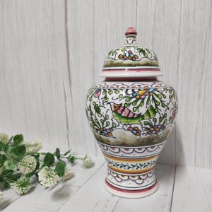 Pode incluir: Um pote de cerâmica branca com tampa, decorado com um padrão floral em tons de verde, rosa e amarelo. O pote tem uma borda festonada e uma base com uma faixa laranja e amarela.