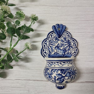 May include: A blue and white ceramic wall planter with a floral design and a rabbit. The planter has a scalloped edge and a small spout at the bottom.