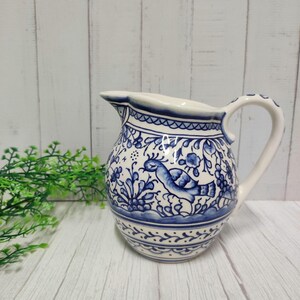 Könnte beinhalten: Weiß-blaue Keramikkrug mit Vogel- und Blumendesign. Der Krug hat einen gebogenen Griff und einen Ausguss zum Ausgießen. Das blau-weiße Muster wiederholt sich am Körper des Kruges.