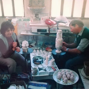May include: Two people are painting ceramic pottery. A woman is painting a white ceramic pot with a blue and green design. A man is painting a large white ceramic vase with a floral design. There are many other ceramic pieces on the table, including small cups and bowls.