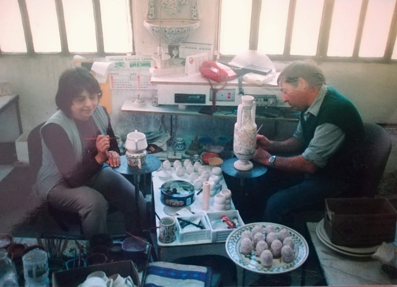 May include: Two people are painting ceramic pottery. A woman is painting a white ceramic pot with a blue and green design. A man is painting a large white ceramic vase with a floral design. There are many other ceramic pieces on the table, including small cups and bowls.