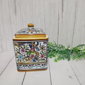 Pode incluir: Um pote quadrado de cerâmica com tampa, apresentando um design floral e de pássaros colorido. O pote é decorado com um fundo amarelo, detalhes brancos e detalhes florais azuis, verdes, rosas e laranja.