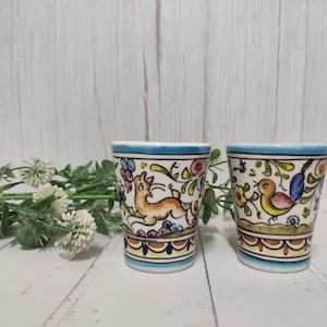 Op de afbeelding: Twee kleine keramische bekers met blauwe en witte randen. De bekers zijn versierd met kleurrijke bloemenpatronen en afbeeldingen van een konijn en een vogel.