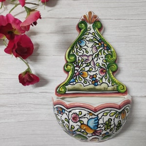 Puede incluir: Un plato de pared de cerámica blanca con un borde rosa y un diseño floral en tonos verdes, azules, rosas y amarillos. El plato tiene un borde festoneado y una pequeña abertura en la parte superior.