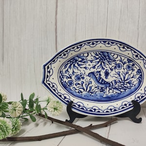 Pode incluir: Um prato de servir oval de cerâmica branca com um design floral azul e branco. O prato apresenta um pássaro e motivos florais. O prato está em um suporte preto.