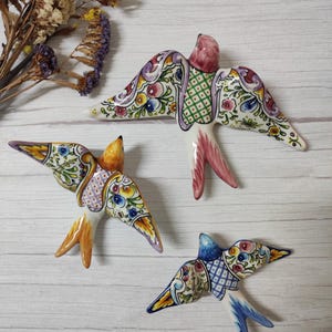 Könnte beinhalten: Drei Schwalben-Wanddekorationen aus Keramik mit handbemalten Blumen- und geometrischen Mustern. Die Vögel sind in Rosa, Gelb und Blau gehalten und verfügen jeweils über eine glänzende Oberfläche und ein markantes, stilisiertes Flügeldesign.