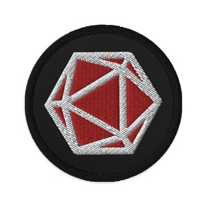 Peut inclure: Un écusson brodé noir et blanc avec un motif géométrique rouge, blanc et noir. L'écusson est circulaire et a une bordure noire.