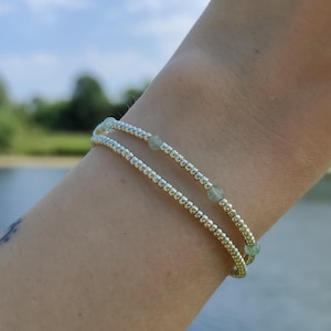 May include: A double-strand bracelet with small, silver-colored beads and interspersed pale green gemstones. The bracelet is worn on a wrist, with a blurred background of water and sky.