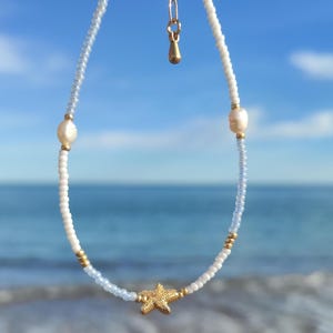 Puede incluir: Una delicada tobillera de cuentas blancas con un dije de estrella de mar dorada y acentos de perlas. La tobillera se sostiene contra un fondo de océano y cielo azul, mostrando su tema playero.