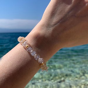 Könnte beinhalten: Ein Armband aus transparenten und goldenen Perlen mit der Aufschrift "LOVE" und goldenen Akzenten. Das Armband wird an einem Handgelenk mit blauem Ozean und Himmel im Hintergrund getragen.